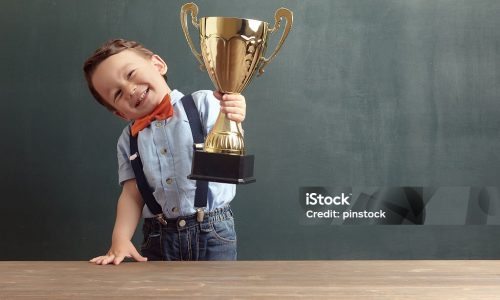 A cute, smiling 2-3 years old boy is standing behind a wooden table and raising a golden trophy with his hand. Little boy is wearing an orange bow tie and blue trousers with suspenders.